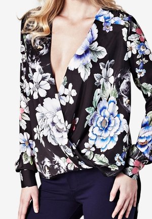 Blouse noire portefeuille avec grands motifs floraux blancs, bleus et roses, portée avec un pantalon foncé, à manches longues et col en V profond.