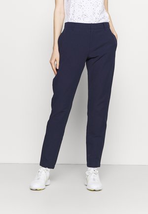 Frau trägt marineblaue, schmal zulaufende Hose, weiß gesprenkeltes Oberteil und weiße Sportschuhe vor einem schlichten hellen Hintergrund.