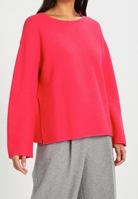 Różowy sweter prążkowany z szerokim dekoltem i długimi rękawami, charakteryzujący się luźnym krojem i bocznymi rozcięciami, w połączeniu z szarymi spodniami.