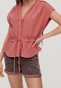 Femme portant un chemisier texturé rose poussiéreux à boutons avec une ceinture à la taille et un ourlet roulé, assorti à un short en denim rose foncé, sur un fond uni.