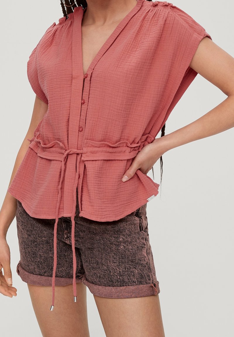 Femme portant un chemisier texturé rose poussiéreux à boutons avec une ceinture à la taille et un ourlet roulé, assorti à un short en denim rose foncé, sur un fond uni.