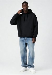 Fekete kapucnis pulóver elülső zsebbel és húzózsinórokkal, világoskék farmernadrággal és fehér tornacipővel párosítva.