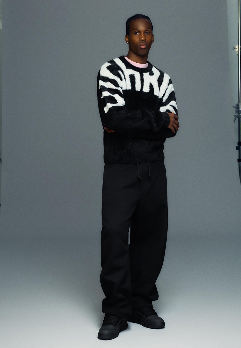 Black fuzzy sweater with white abstract pattern, paired with loose black pants and black sneakers. Model poses with arms crossed.