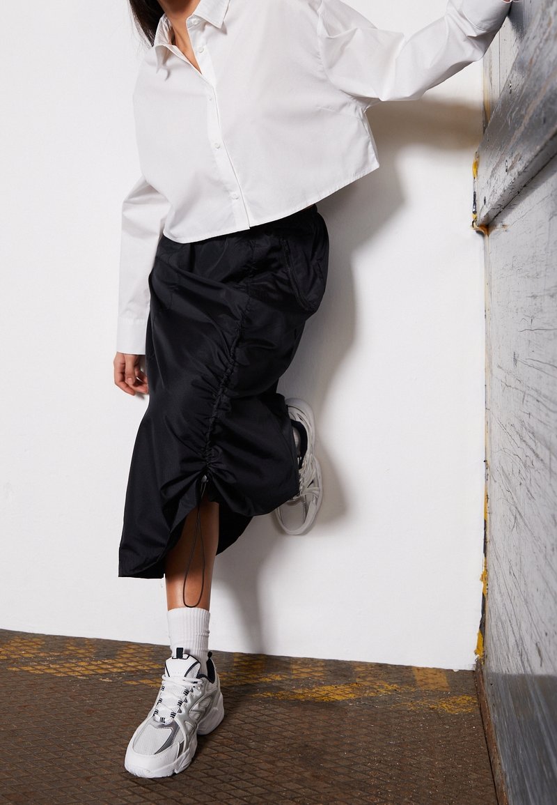 Personne portant un haut court blanc, une jupe midi noire froncée, des chaussettes blanches et des baskets blanches, appuyée contre un mur avec un pied levé.