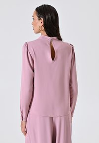 Blusa a maniche lunghe rosa chiaro con apertura a chiave sul retro, chiusura a bottone e dettagli arricciati sulle spalle. Tessuto morbido e fluido.