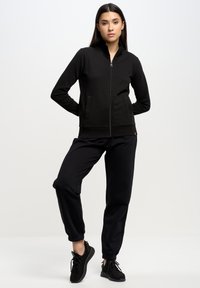 Melnas zip-up jaka ar garām piedurknēm un sānu kabatām, apvienota ar saskanīgiem melniem jogger biksēm un melniem sporta apaviem. Mīksts auduma faktūra.