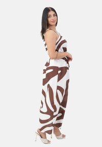 Kvinde med langt mørkt hår iført en ærmeløs hvid jumpsuit med brune abstrakte mønstre og hvide højhælede sandaler, stående sidelæns.