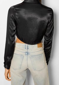 Svart satin cropped skjorta med långa ärmar, krage och knäppning vid ärmsluten, matchad med ljusblå-högmidjade denimjeans.