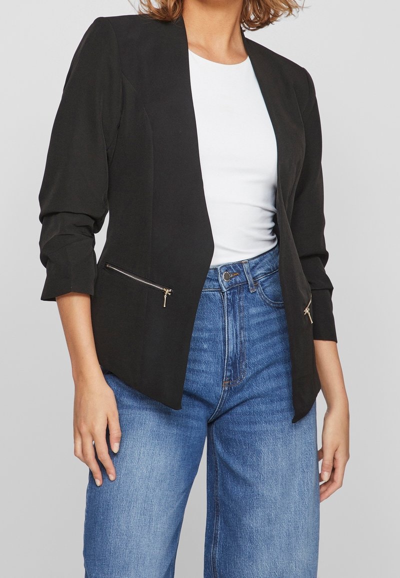 Femme portant un blazer noir avec poches zippées, un top blanc et un jean bleu taille haute sur fond uni.