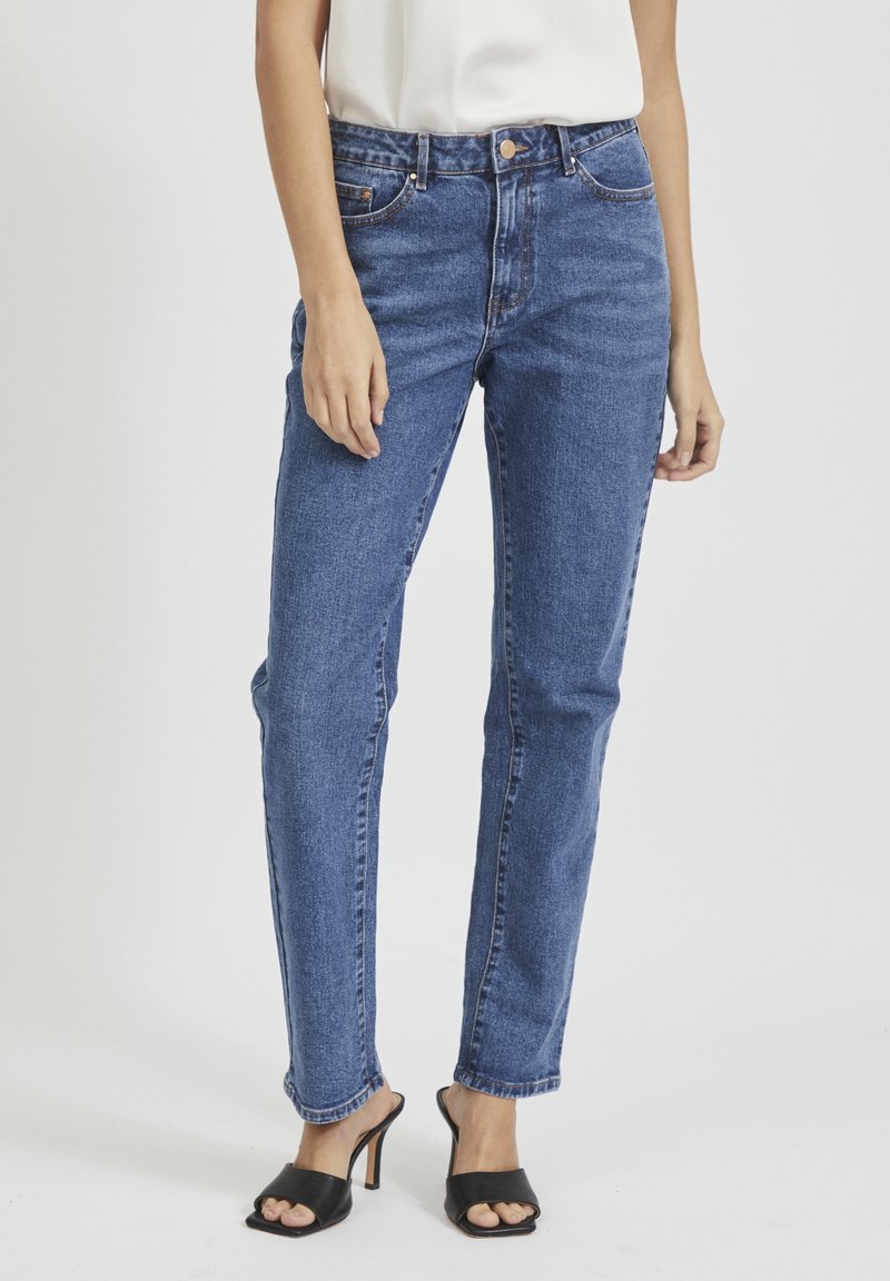 Femme portant un jean bleu taille moyenne à jambes droites, un haut blanc et des sandales à talons noires à bouts ouverts, debout devant un fond blanc uni.