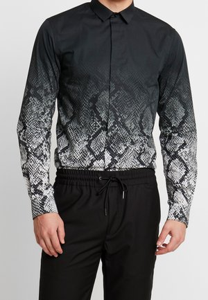 Mand iført en sort skjorte med knapper med hvidt og gråt slangeskindsmønster på ærmer og krop, kombineret med sorte bukser med snøre.