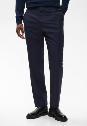 Homme portant un pantalon bleu marine foncé sur mesure et des bottes en cuir noir, debout avec une main dans la poche devant un fond uni.