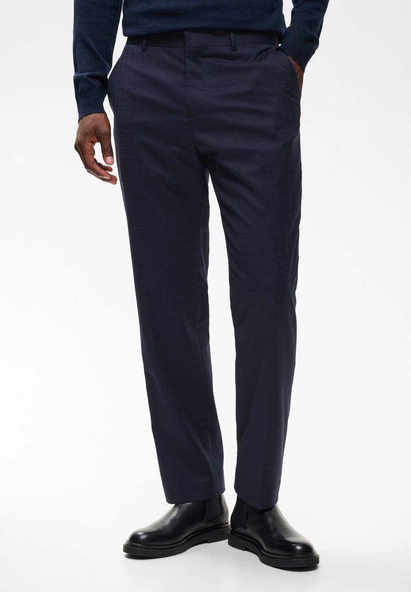 Homme portant un pantalon bleu marine foncé sur mesure et des bottes en cuir noir, debout avec une main dans la poche devant un fond uni.