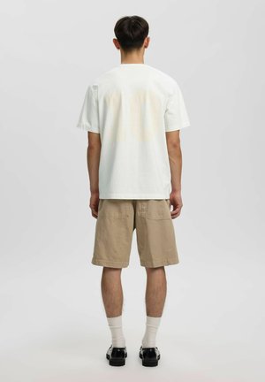 Homme debout, dos à la caméra, portant un t-shirt blanc uni, un short beige au genou, des chaussettes blanches et des chaussures noires sur un fond uni.