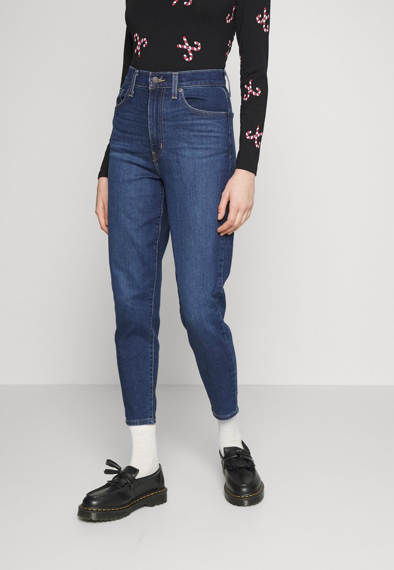 Jeans en denim bleu taille haute avec une coupe fuselée. Le tissu semble doux avec des détails de couture subtils. Portés avec des mocassins noirs et des chaussettes blanches.