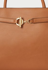 Torebka skórzana w kolorze tan, o gładkiej teksturze, z okrągłymi metalowymi detalami w odcieniu złotym oraz płaskim paskiem. Minimalistyczny design, czyste linie.