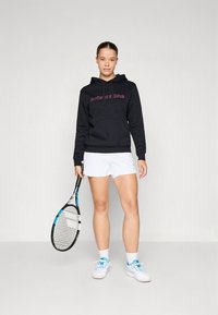 Camisola preta com capuz e logótipo rosa, calções brancos e a segurar uma raquete de ténis. O conjunto inclui sapatilhas brancas com detalhes azuis.