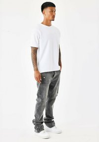 Camiseta blanca de manga corta con cuello redondo, combinada con jeans grises desgastados y zapatillas blancas. Modelo de pie en un fondo neutro.
