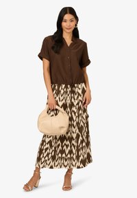 Chemise cintrée à manches courtes de couleur brune, associée à une jupe maxi fluide à motifs dans des teintes de brun et de crème. Tenant un sac à main beige.