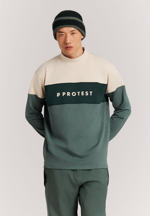 Young man wearing a green and beige color-block sweater with "PROTEST" logo and a dark striped knit beanie, standing with hands behind back.
