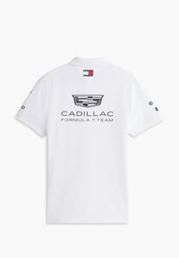 Λευκό πόλο μπλουζάκι με το λογότυπο της Cadillac και το κείμενο "Formula 1 Team" στην πλάτη και μια μικρή σημαία Tommy Hilfiger κάτω από το γιακά.