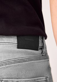 Blugi gri din denim cu o etichetă neagră texturată pe banda din talie. Materialul prezintă un model subtil de țesătură diagonală.