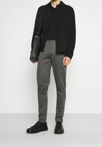 Man wearing black jacket, black shirt, gray pants, black shoes, carrying black shoulder bag against white background.