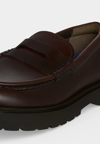 Gros plan sur un mocassin en cuir marron foncé avec une épaisse semelle en caoutchouc noir et une bride fendue sur le dessus.