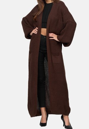 Femme portant un long cardigan marron en maille avec des manches bouffantes, un top court noir, un pantalon noir et des talons hauts noirs, debout les mains sur les hanches.