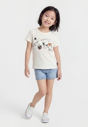 Glimlachend kind met kort zwart haar draagt een crèmekleurig T-shirt met cartoonfiguren, lichte denimshorts en witte sneakers, staand op een witte achtergrond.