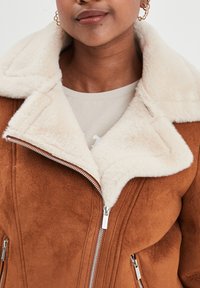 Braune Fake-Suede-Jacke mit einem krämfarbenen Kunstfellkragen, ausgestattet mit silbernen Reißverschlüssen und einer strukturierten Oberfläche.