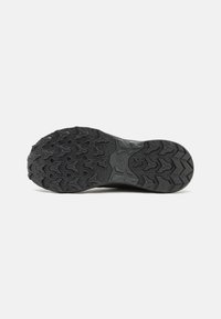 Suela de zapatilla de trail negra con un patrón de tracción profunda, que presenta tacos prominentes y formas geométricas variadas para un agarre en diferentes terrenos.