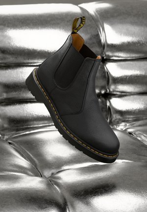 Black leather Chelsea boot with elastic side panels, yellow stitching, and a pull tab; displayed on a shiny metallic surface.
