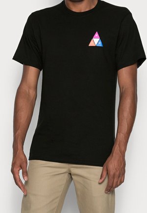 Homme portant un t-shirt noir avec un petit logo triangulaire multicolore sur la poitrine gauche, associé à un pantalon beige.
