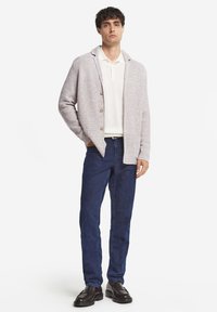 Jasnoszara dziergana kamizelka z kołnierzykiem i drewnianymi guzikami, w połączeniu z białą koszulą polo i ciemnoniebieskimi dżinsami. Czarne buty wsuwane dopełniają całość.