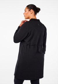 Melnā garā piedurknē mēteli ar apkakli, aizmugurējo krokot un elastīgu jostas detaļu. Izgatavots no mīksta auduma ar gludu tekstūru un minimālistisku dizainu.