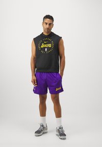 Melnā bezpiedurkņu kapuci ar dzelteno "Lakers" tekstu, apvienota ar purpura sieta šortiem, kuros redzams "Lakers" teksts, pelēkiem sporta apaviem un baltām zeķēm.