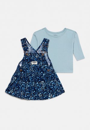 Chemise bleue à manches longues associée à une robe chasuble bleu marine à imprimé floral, avec des bretelles, des boutons et une jupe évasée.