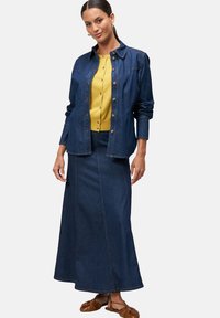 Veste en denim avec fermeture à boutons, associée à un haut en tricot jaune et une longue jupe en denim. Des chaussures plates marron complètent l'ensemble.