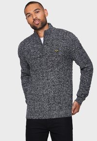 Strikket sort og hvid sweater med høj krave og halvdels lynlås, med et gult logoaccent på brystet. Struktureret, varmt stof.