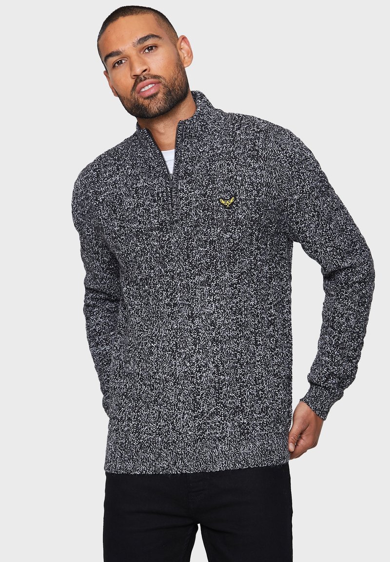 Strikket sort og hvid sweater med høj krave og halvdels lynlås, med et gult logoaccent på brystet. Struktureret, varmt stof.