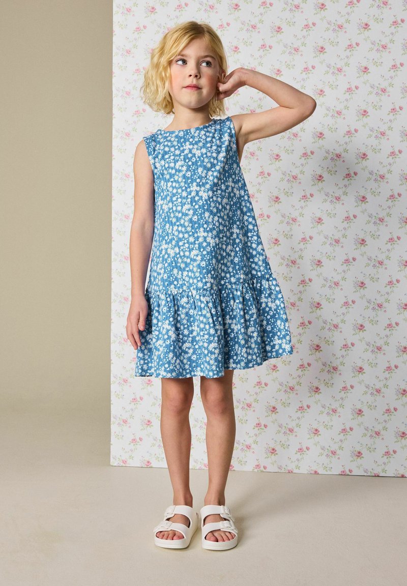 Robe à fleurs bleues avec un ourlet évasé, fabriquée en tissu respirant. Portée avec des sandales blanches. Le fond présente un motif floral.