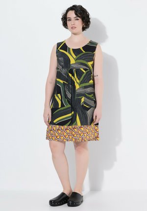 Femme aux cheveux courts et bouclés portant une robe sans manches à motifs avec des sabots noirs, debout devant un fond blanc.