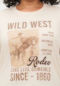 Krēma krāsas kokvilnas T-krekls ar vintage rodeo grafiku brūnos toņos, ar izteiksmīgu tekstu "WILD WEST" un "RODEO" dažādos fontos.