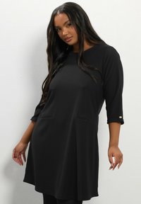 Tunic nera con maniche a tre quarti, caratterizzata da due tasche frontali e dettagli di bottoni sulle maniche. Texture morbida e vestibilità rilassata.