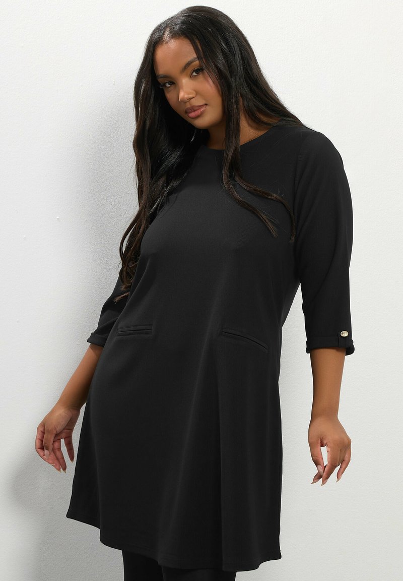 Tunic nera con maniche a tre quarti, caratterizzata da due tasche frontali e dettagli di bottoni sulle maniche. Texture morbida e vestibilità rilassata.