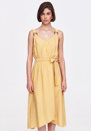 Sukienka do kolan w żółtym kolorze, wykonana z miękkiego materiału, z dekoltem w kształcie litery V, szerokimi ramiączkami z okrągłym metalowym elementem orazwiązanym paskiem w talii.