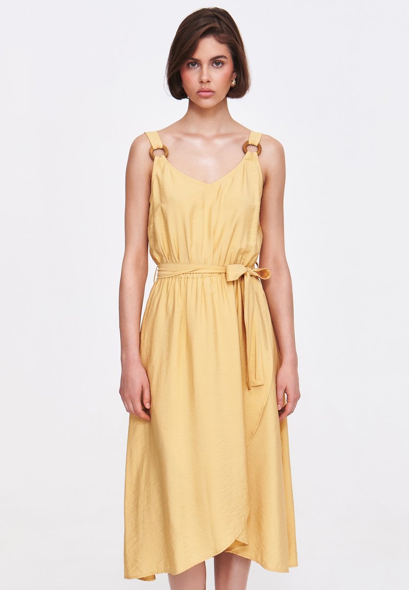 Sukienka do kolan w żółtym kolorze, wykonana z miękkiego materiału, z dekoltem w kształcie litery V, szerokimi ramiączkami z okrągłym metalowym elementem orazwiązanym paskiem w talii.