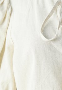 Tessuto color crema con un aspetto testurizzato simile al lino. Presenta dettagli arricciati e una chiusura con fascetta. Materiale morbido e leggero.
