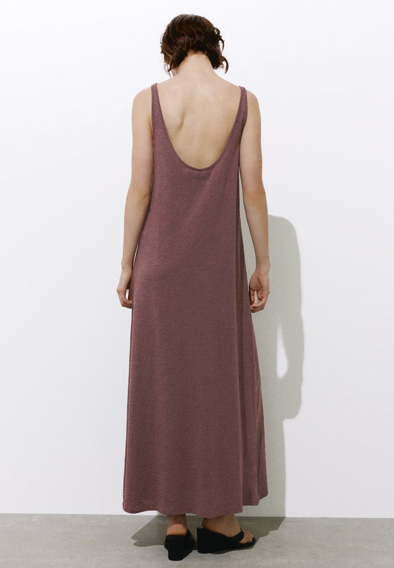Personne aux cheveux courts et bouclés portant une robe longue mauve sans manches avec un dos décolleté et des sandales compensées noires, debout contre un mur blanc.
